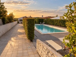 Una piscina rettangolare con bordo piastrellato, circondata da un basso muro in pietra e un siepe. Sono presenti sedie a sdraio e un tavolo vicino alla piscina.