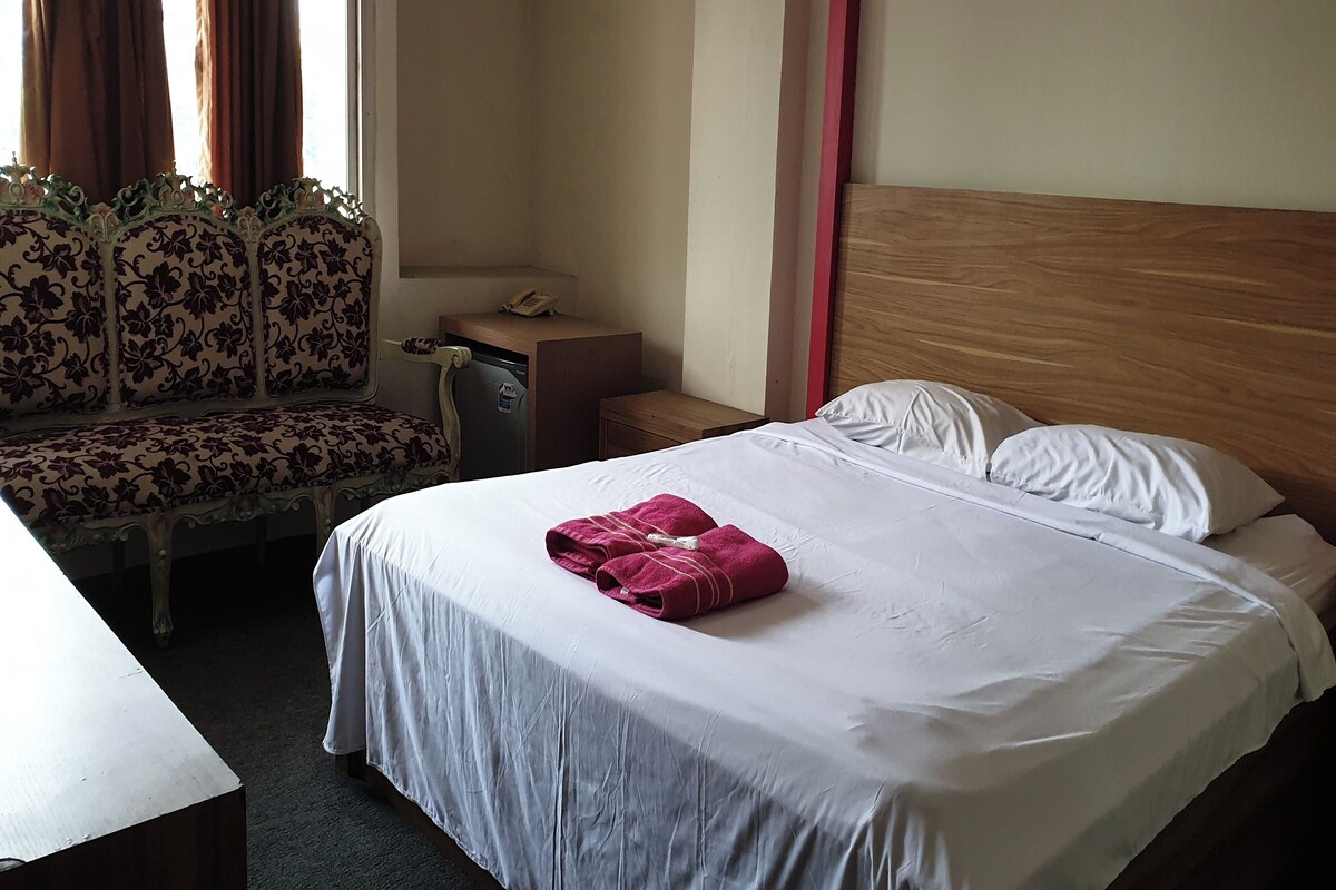Une chambre d’hôtel avec un lit, une tête de lit en bois, un banc recouvert d’un tissu à motifs floraux et un téléphone posé sur une petite table.