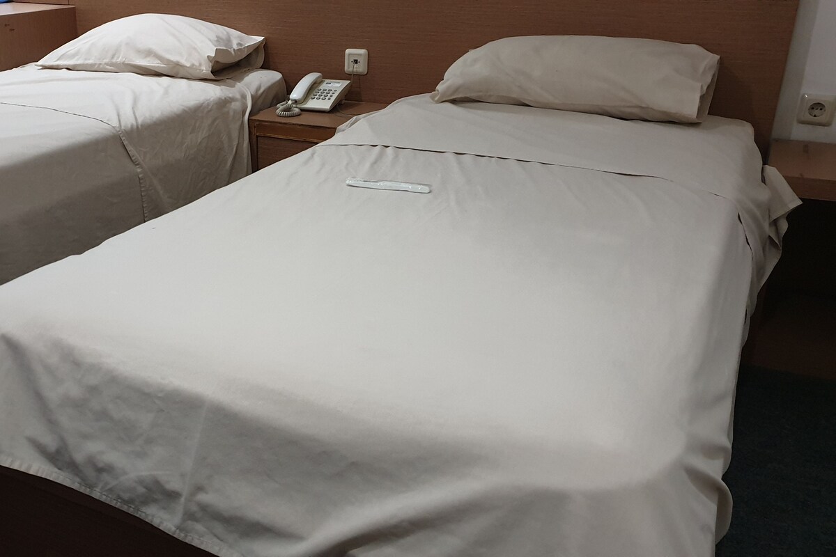 Deux lits simples avec du linge de lit blanc, une table de chevet avec un téléphone et une lampe, et une prise électrique au mur.