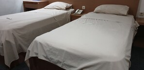 Deux lits simples avec du linge de lit blanc, une table de chevet avec un téléphone et une lampe, et une prise électrique au mur.