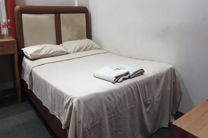 Un lit bien fait, recouvert d’une couvre-lit beige et agrémenté d’oreillers, avec une tête de lit en bois et une petite table sur laquelle sont posées des bouteilles.