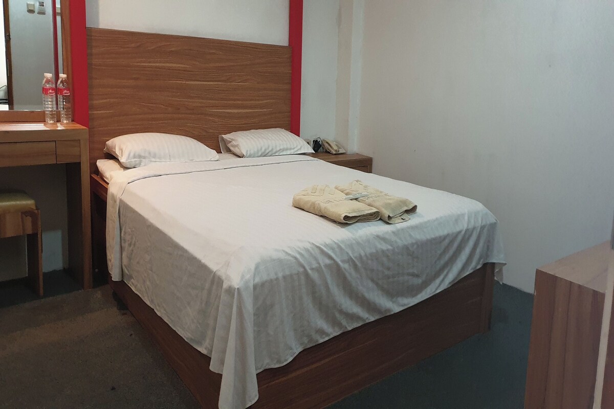 Un lit bien fait, recouvert d’une couvre-lit blanche et agrémenté de deux oreillers, une tête de lit en bois, une petite table de chevet avec un téléphone, et un bureau avec une chaise et deux bouteilles d’eau.