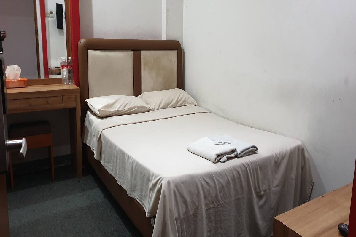 Una cama bien hecha con cubrecama y almohadas blancas, una mesita de noche de madera con un dispensador de pañuelos y una botella, y un escritorio de madera con un espejo.