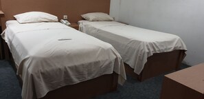 Deux lits simples avec du linge de lit blanc, un téléphone sur la table de chevet et une petite table dans une chambre d’hôtel.