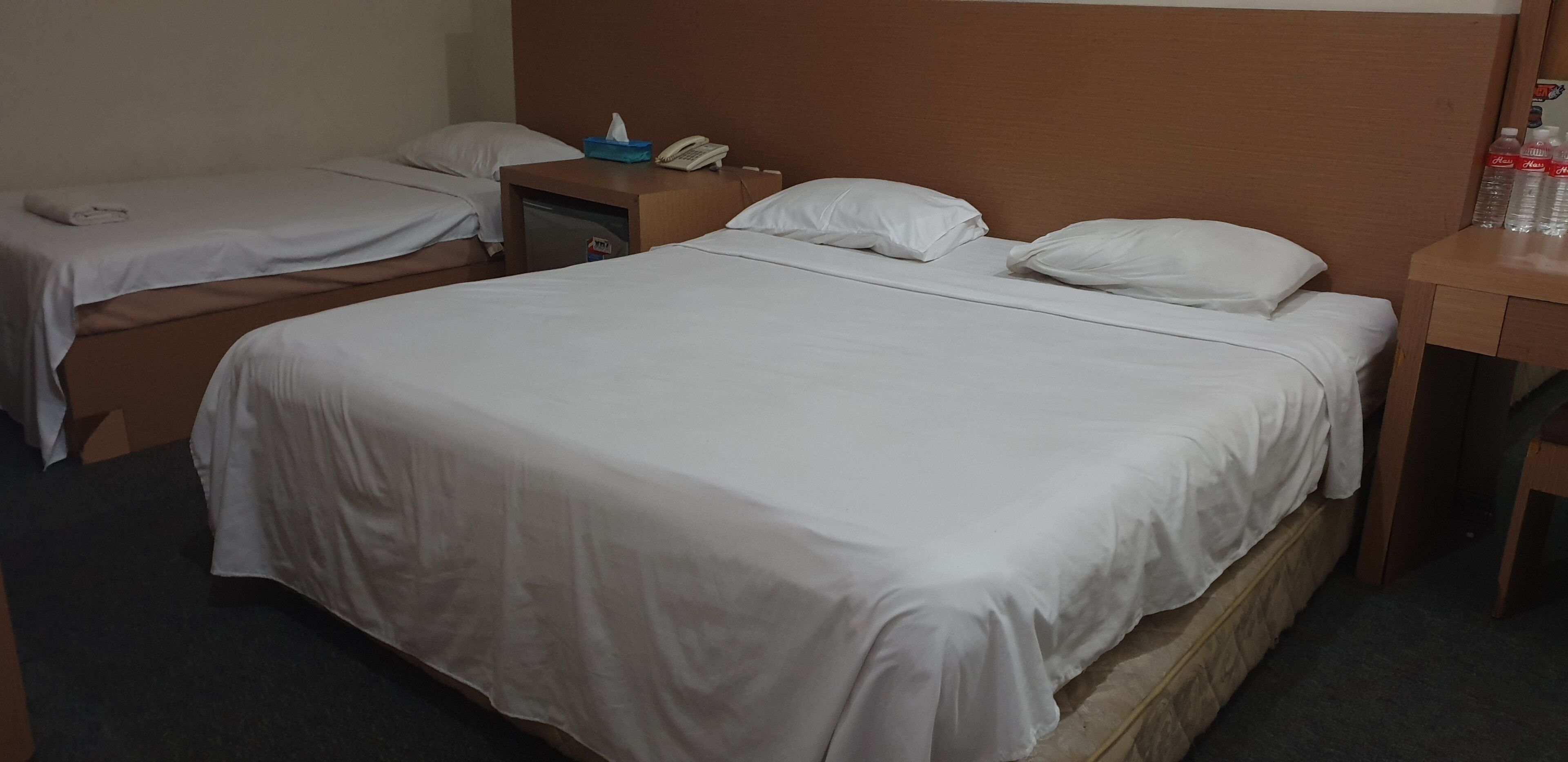 Habitación de hotel con dos camas individuales, cabecera de madera, mesita de noche con teléfono y pañuelos, y una pequeña mesa con botellas de agua.