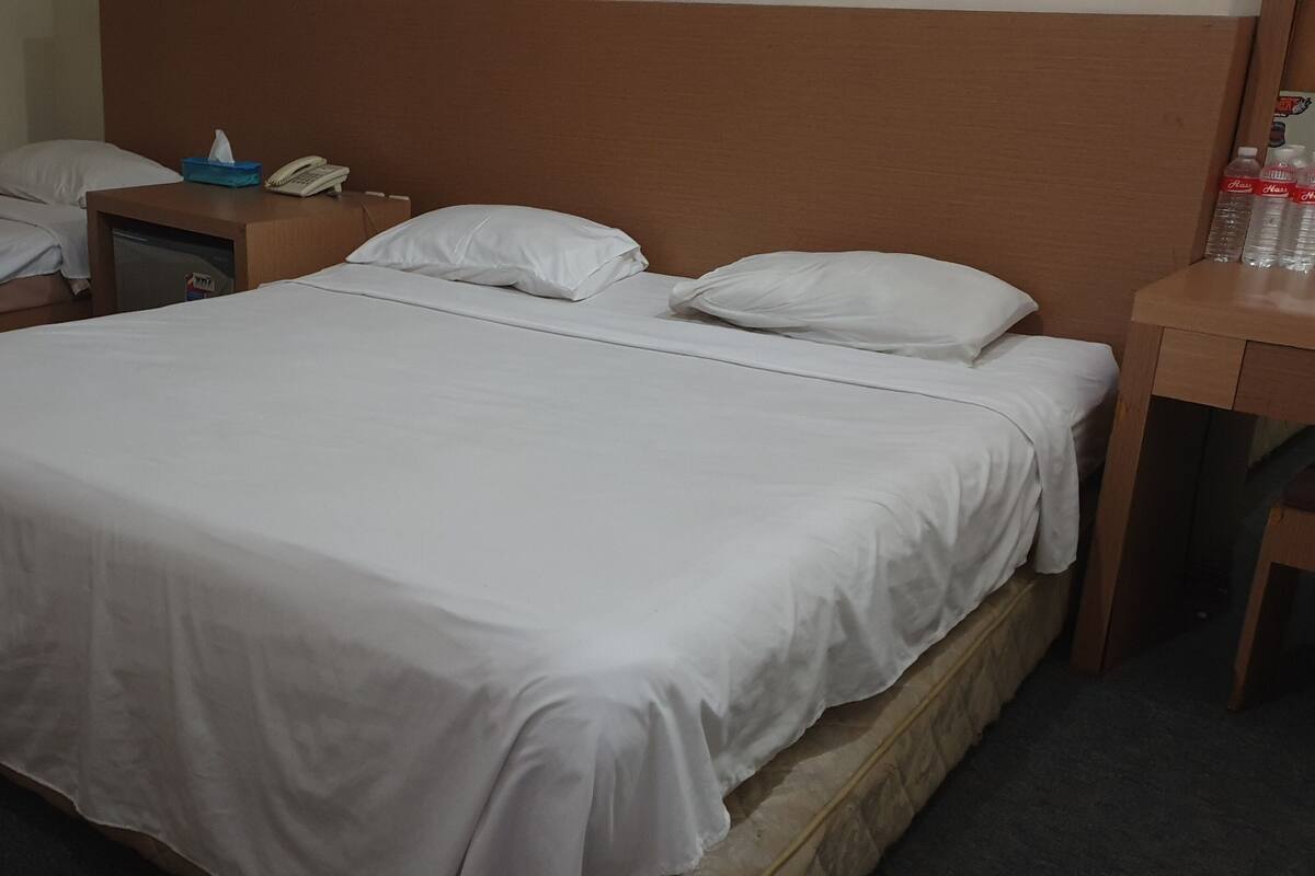 Habitación de hotel con dos camas individuales, cabecera de madera, mesita de noche con teléfono y pañuelos, y una pequeña mesa con botellas de agua.