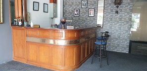 Une zone d’accueil avec un comptoir en bois, une réceptionniste et une caissière. Des tableaux encadrés ornent le mur, et il y a une horloge.