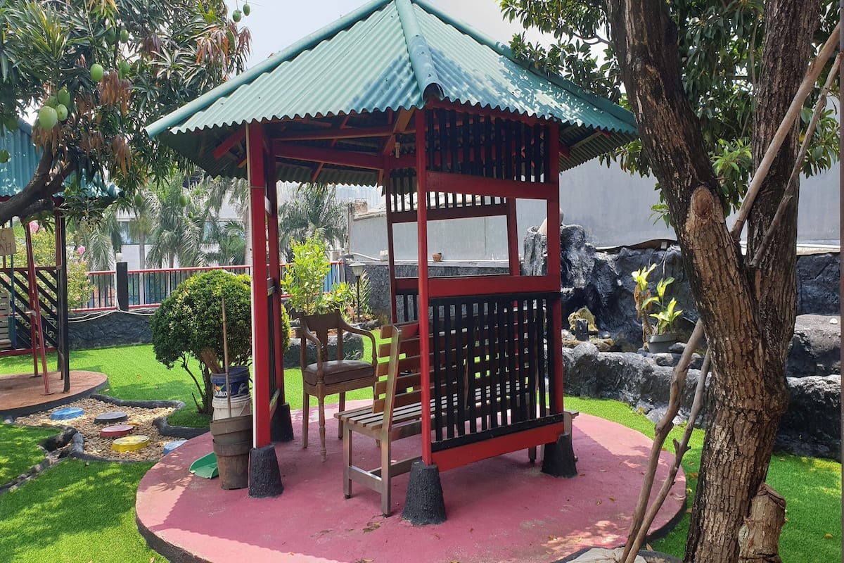 Un pabellón con techo verde y estructura roja, rodeado de un jardín pequeño que incluye una resbaladilla y bancos.