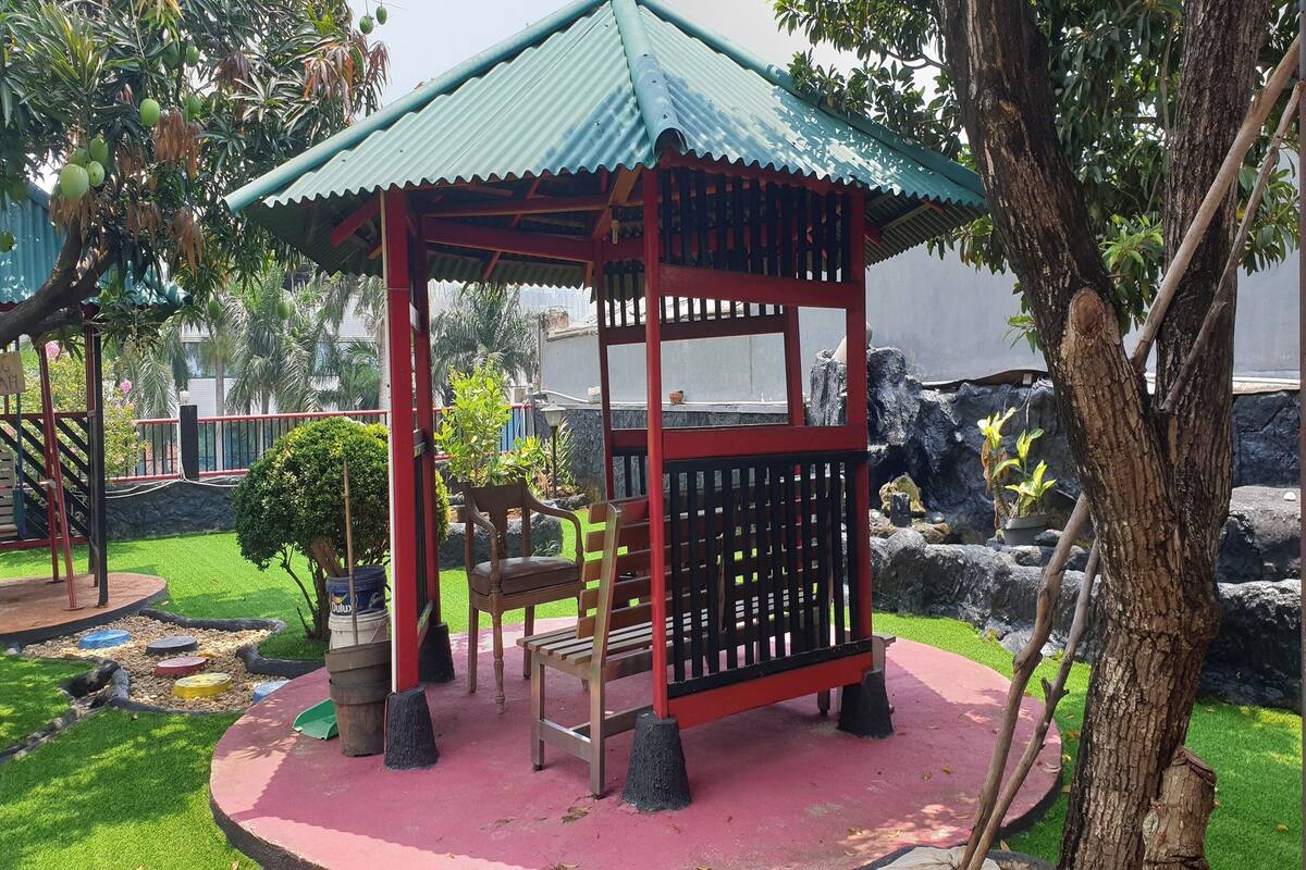 A gazebo with a green roof and red frame, surrounded by a small garden with a swing set and benches.