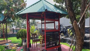 Un kiosque doté d’un toit vert et d’une structure rouge, entouré d’un petit jardin avec une balançoire et des bancs.