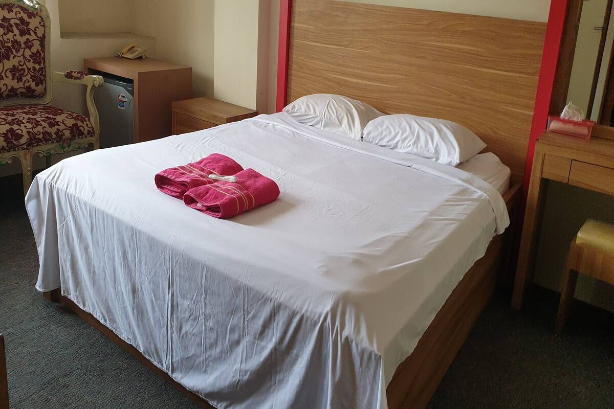 Una cama bien hecha con sábanas blancas y una toalla rosa, una mesita de noche de madera con un teléfono, una silla con estampado floral y una pequeña mesa con un borde rojo.