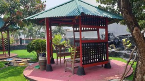 Un kiosque doté d’un toit rouge et vert, entouré d’un jardin où se trouvent un banc et une petite table.