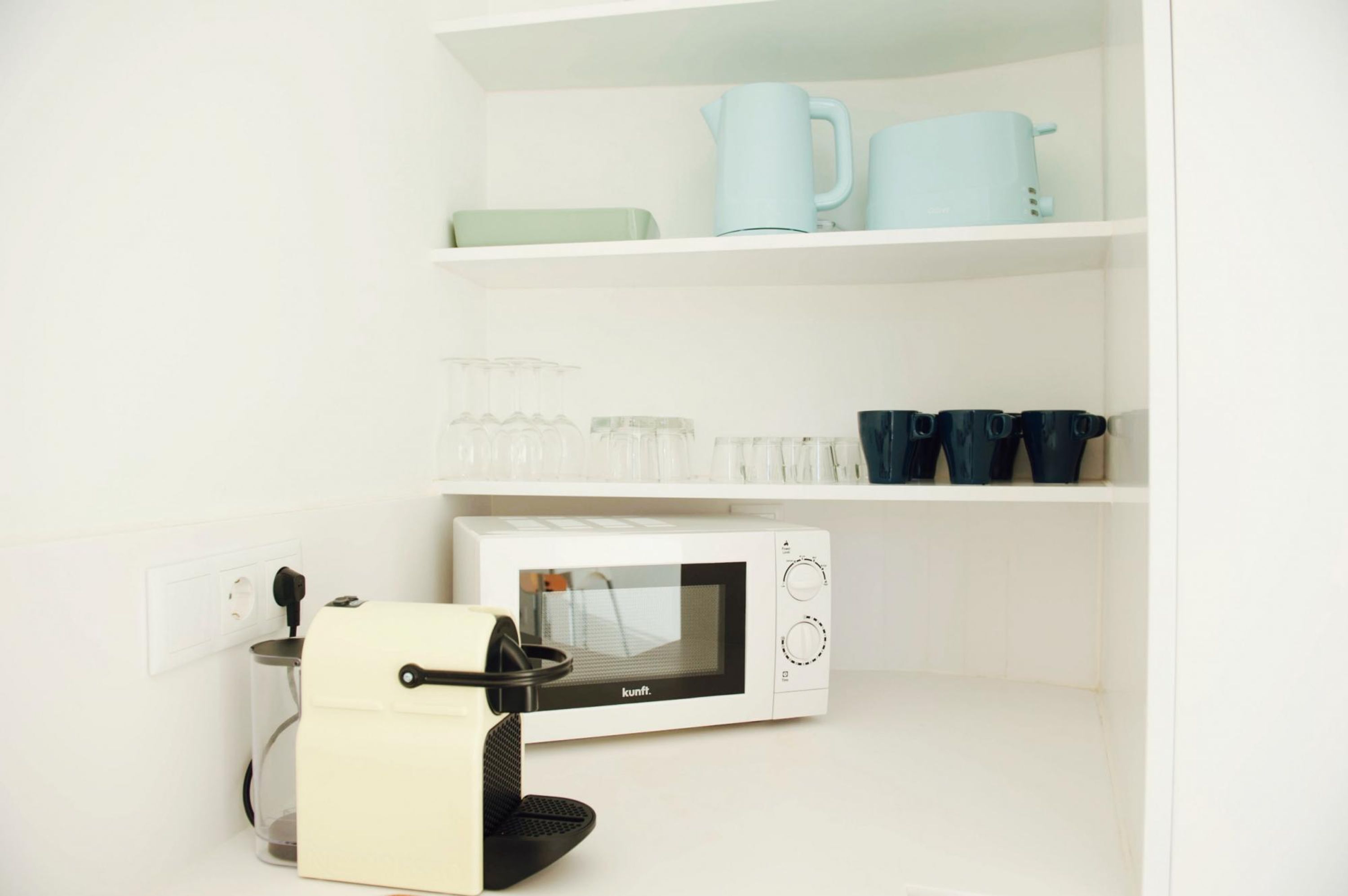 Una cocina equipada con microondas, cafetera y una alacena con tazas y hervidor.