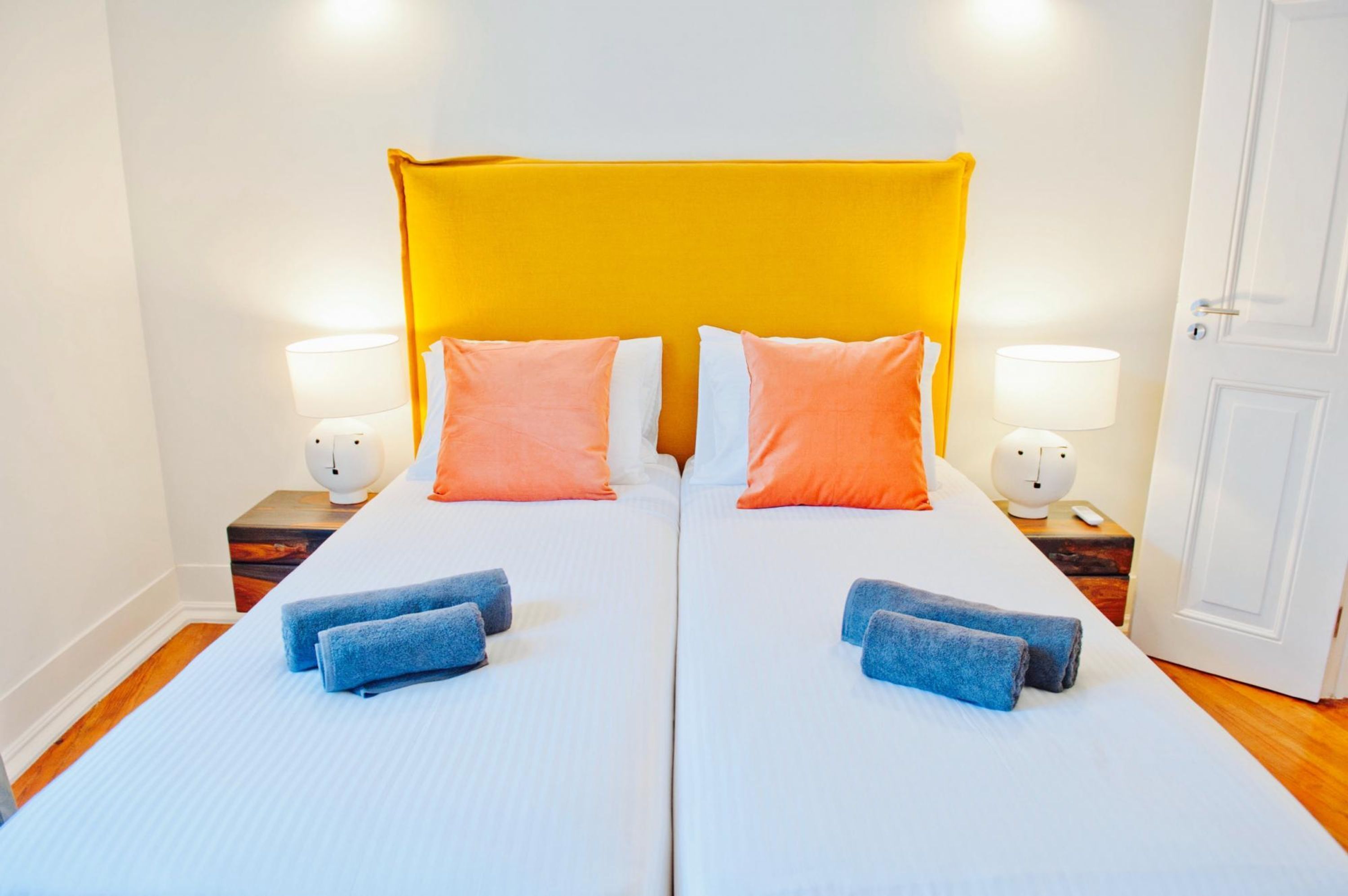 Una cama doble con cabecera amarilla, dos almohadas anaranjadas y dos toallas azules.