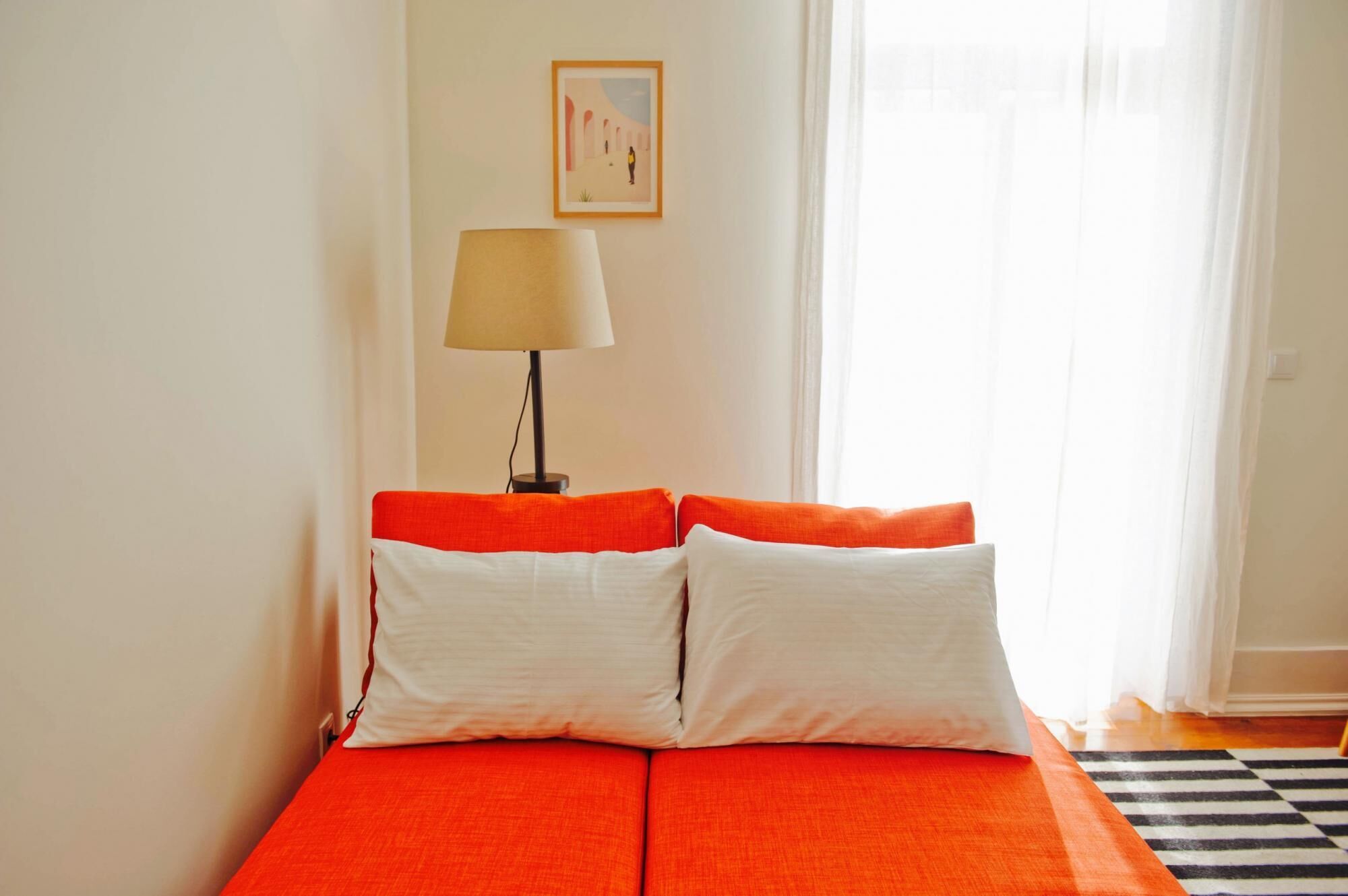 Una cama con dos almohadas blancas y una colcha naranja en una habitación con una lámpara y un cuadro en la pared.