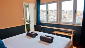 A hotel room with a bed, a wooden bench, a window with a city view, and a door. - Expresso Martinis in Lisbon by Misha s Place 4c (Lisbon)