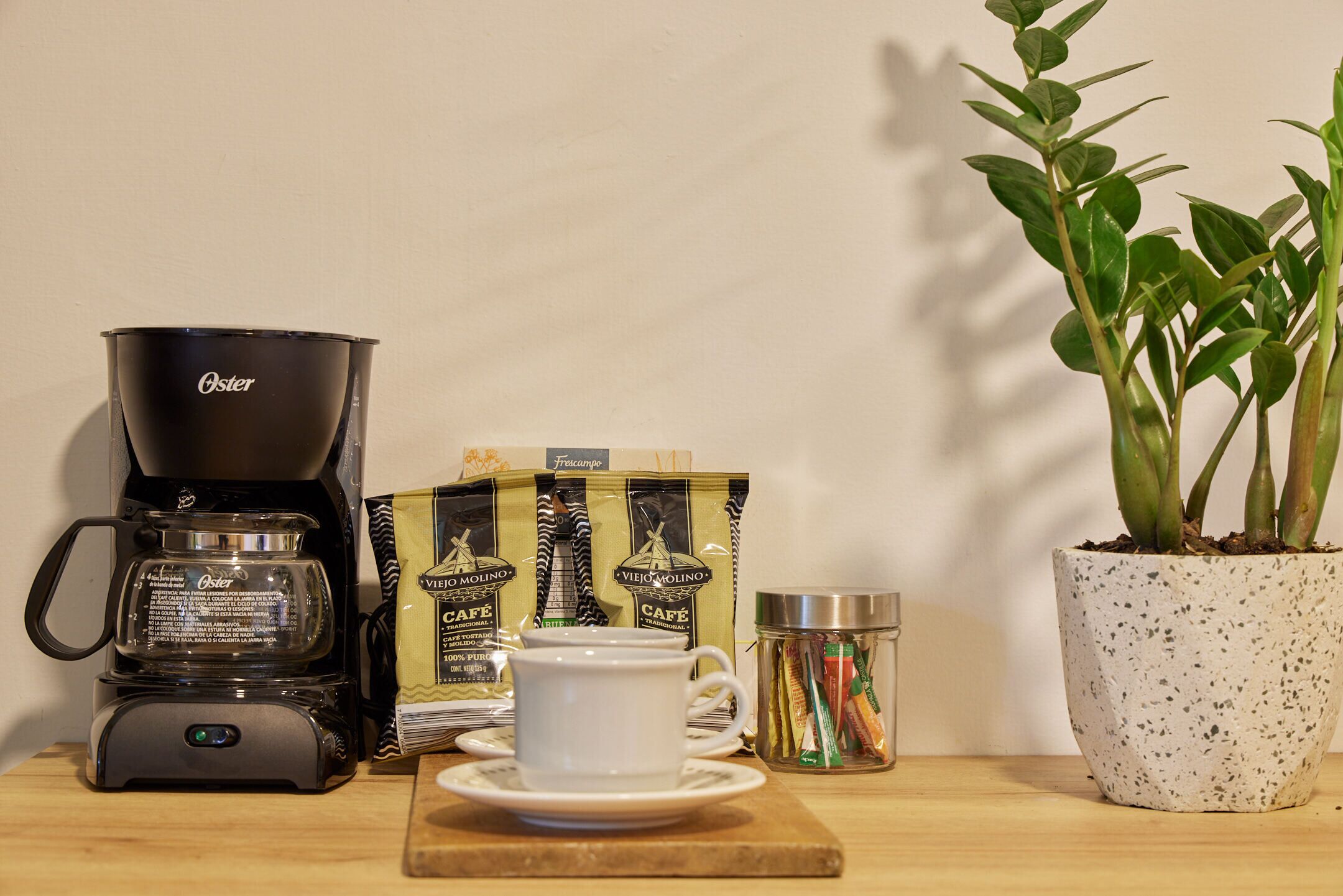 A coffee maker, a cup, a box of coffee, a jar of coffee pods, and a potted plant on a wooden surface.