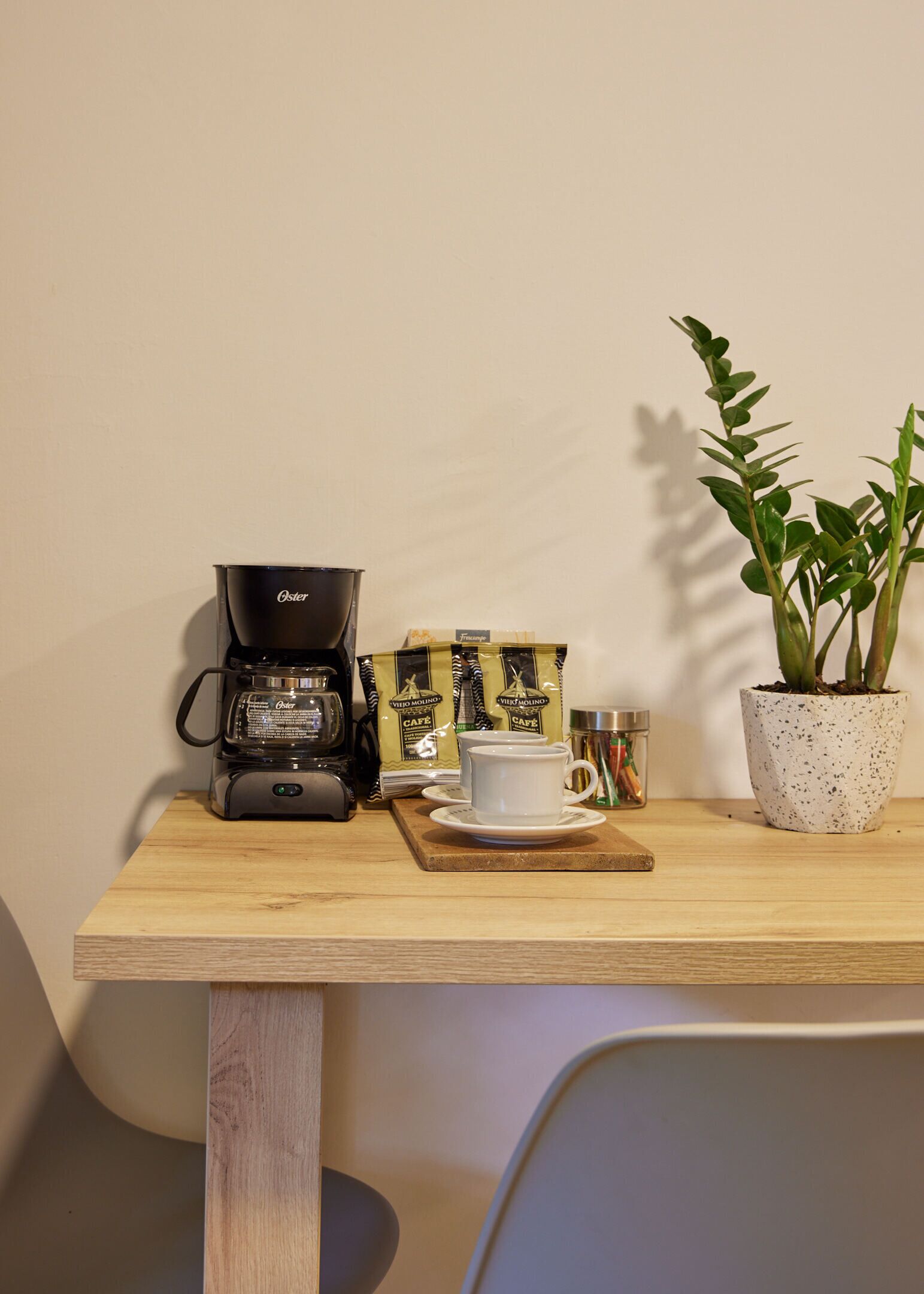 A wooden table with a coffee maker, a white cup, a plant, and coffee packets.