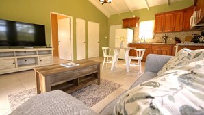 Ein Wohnzimmer mit Fernseher, Couch und Couchtisch. Dazu gehört eine Küchenzeile mit Holzausbau und ein Esstisch mit Stühlen.