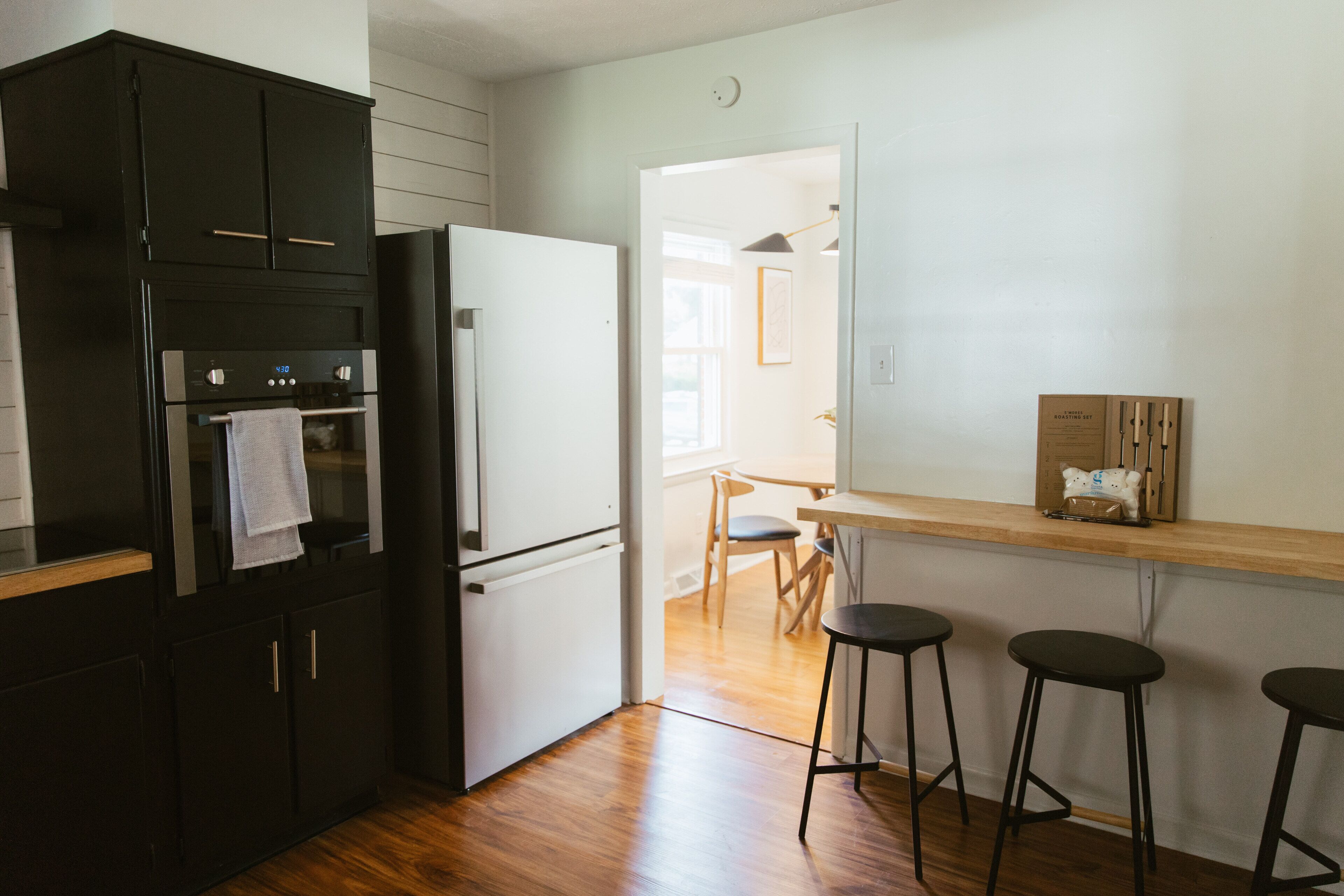 A kitchen with a black refrigerator, a white oven, and wooden flooring. There are three black stools and a wooden bar counter.