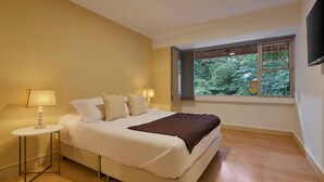 A bedroom with a bed, two bedside lamps, a window with a view of greenery, and a flat-screen TV mounted on the wall. - Provenza's Retreat - Tranquil 2BR-2BA Apartment (Medellín)