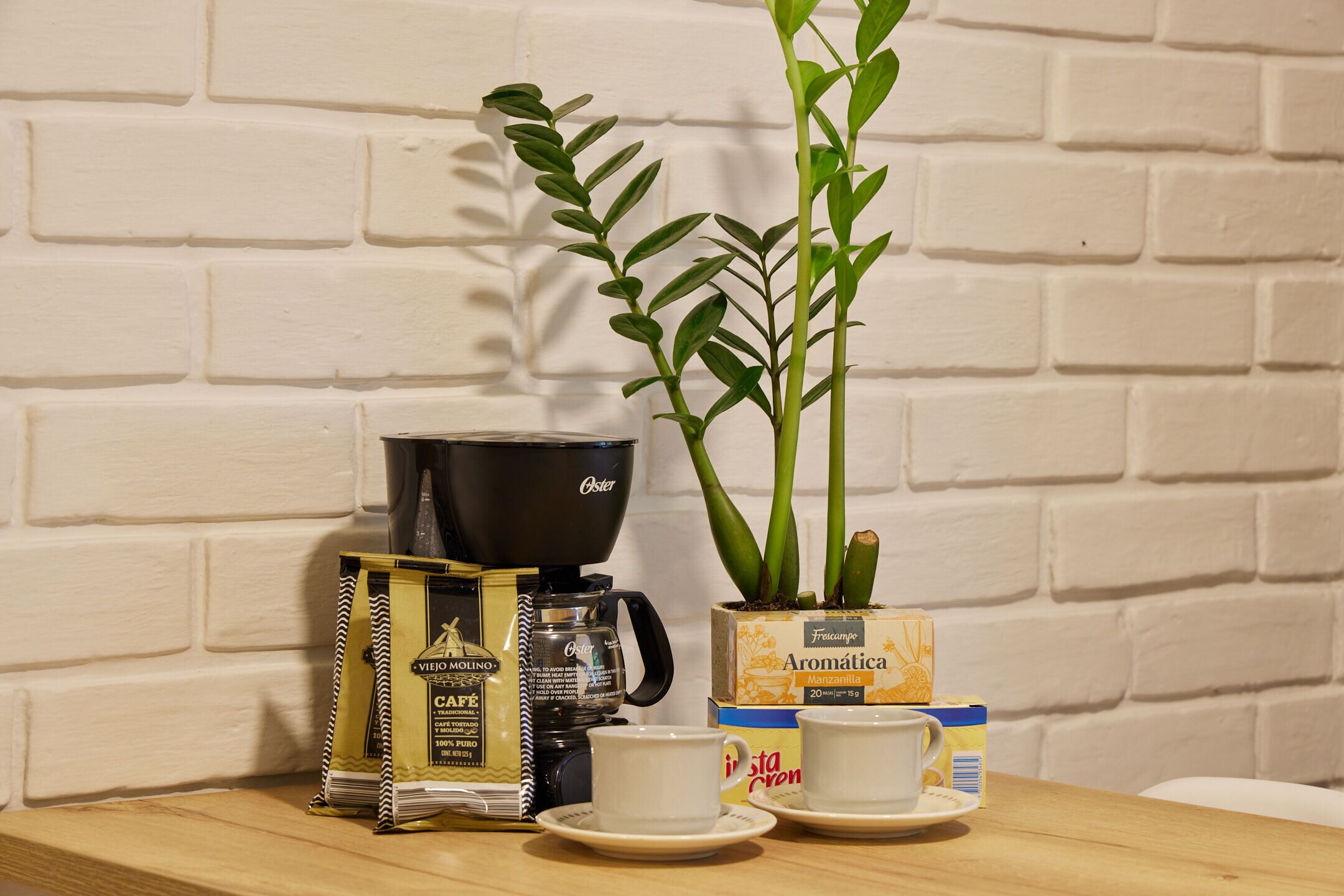 En kaffebryggare, en krukväxt och kaffekapslar står på ett bord framför en vit tegelvägg.