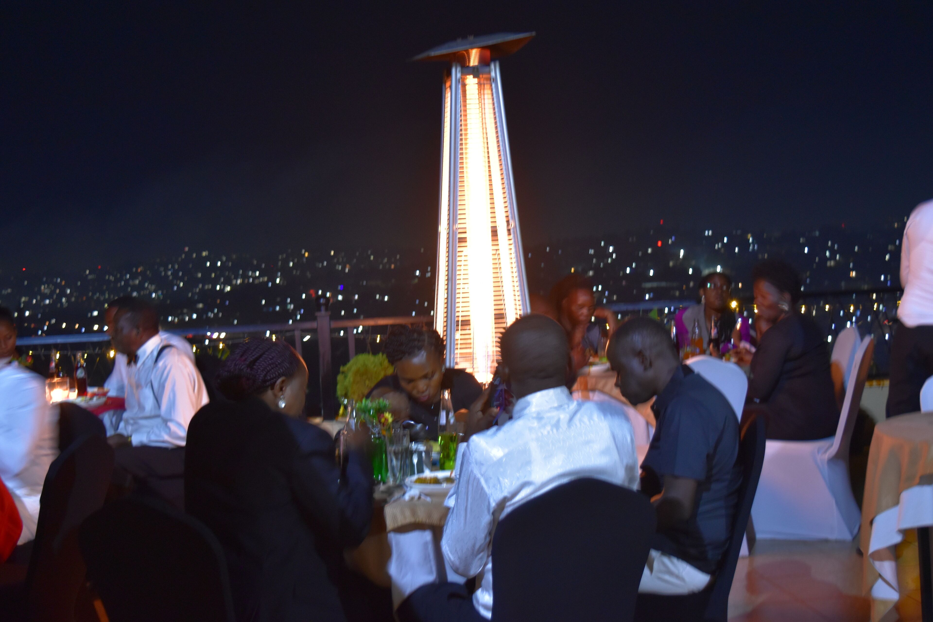 A rooftop dining area with guests seated at tables, a central decorative light, and a cityscape view in the background.