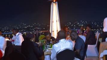 A rooftop dining area with guests seated at tables, a central decorative light, and a cityscape view in the background.