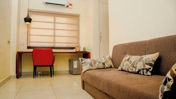 A small, furnished room with a brown sofa, a red chair, a desk, and a window with blinds.