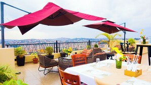 An outdoor dining area with tables, chairs, and umbrellas, overlooking a cityscape. - Millennium Terrace Hotel (Kampala)