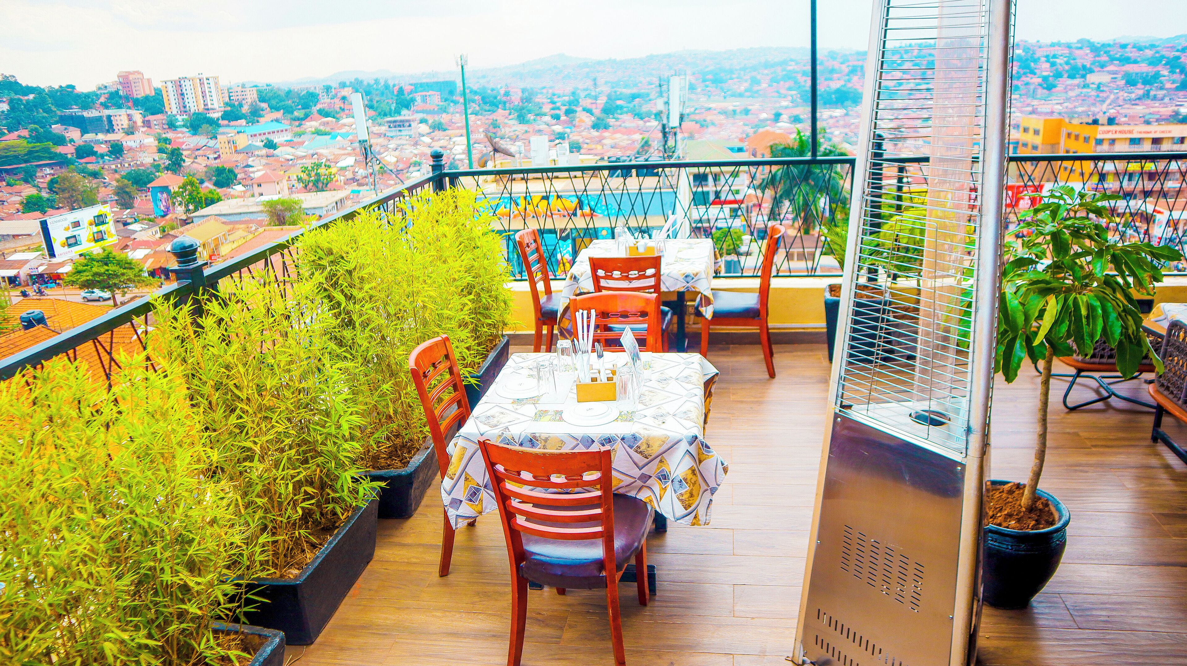 A rooftop restaurant with tables and chairs, surrounded by potted plants and overlooking a cityscape.