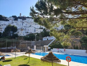 Une piscine équipée d’une bouée de sauvetage, un espace verdoyant agrémenté de parasols en chaume, et une vue sur des bâtiments blancs situés sur une colline.