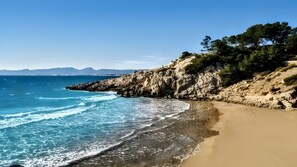 A coastal beach with clear blue water, sandy shore, rocky cliffs, and greenery.