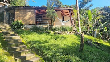 Una casa con una escalera de piedra que lleva a una terraza de madera, rodeada de exuberante vegetación y un cielo azul despejado.