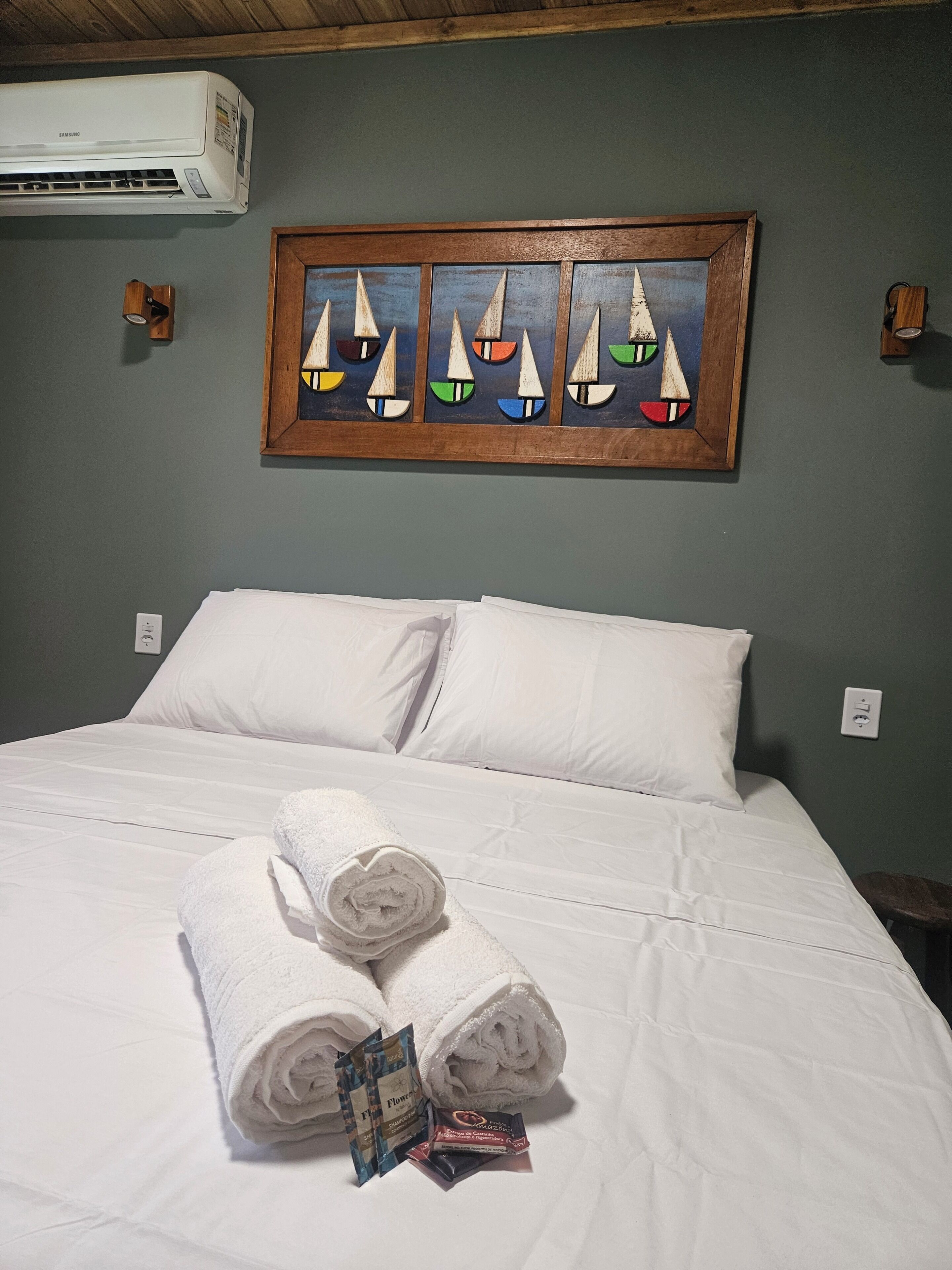 A hotel room with a bed, white linens, a painting of sailboats, and a wall-mounted air conditioner.