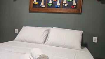 A hotel room with a bed, white linens, a painting of sailboats, and a wall-mounted air conditioner.