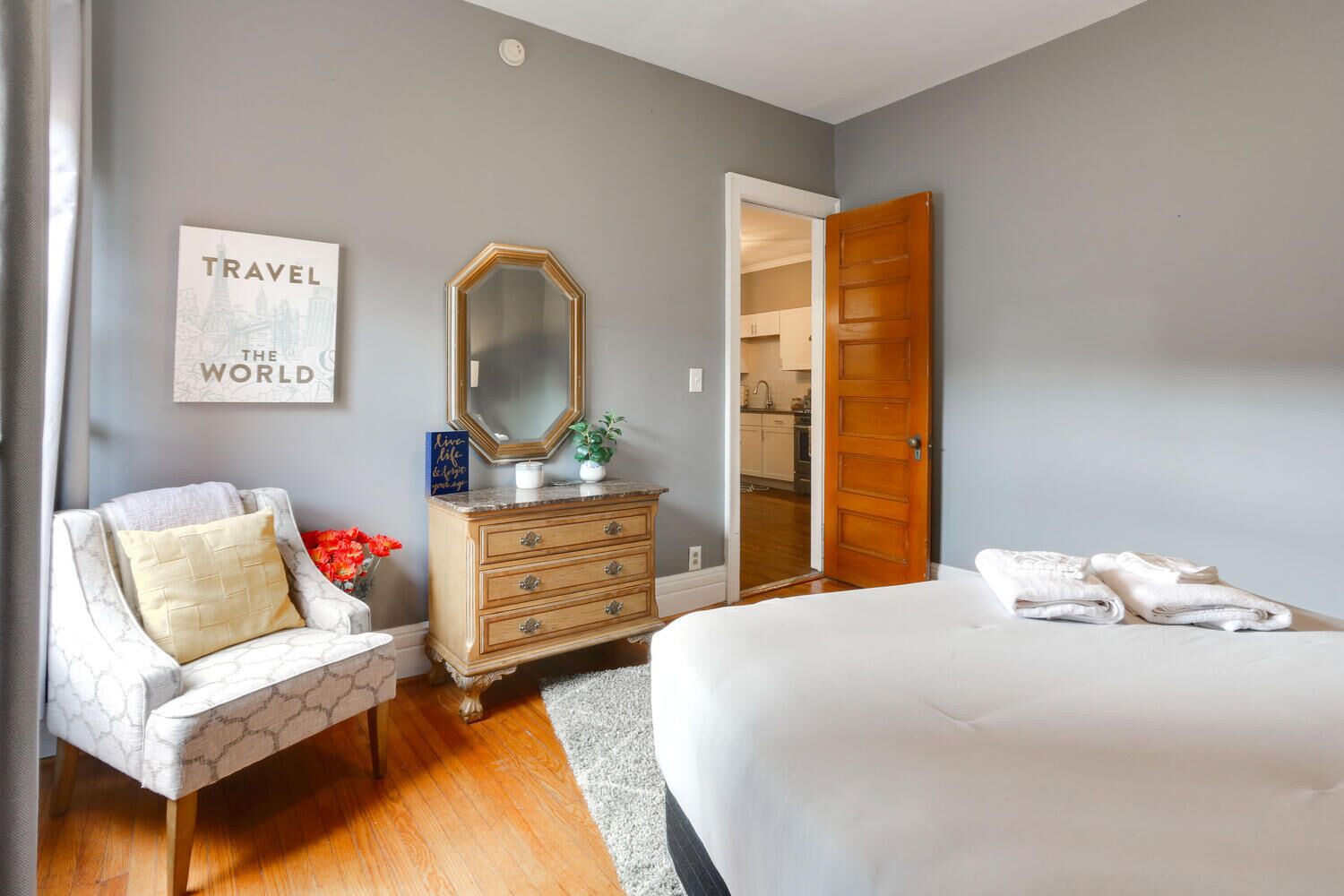 Un dormitorio con una cama, un aparador, una silla, un espejo y una pared decorada con un cartel de temática viajera.