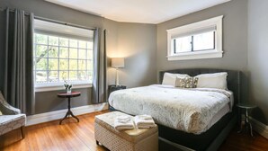 Ein Schlafzimmer mit einem großen Bett, einem Nachttisch, einer Lampe, einem Sessel und einem Fenster mit Vorhängen.