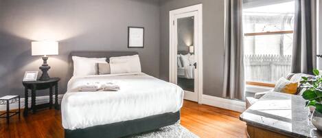 A bedroom with a bed, bedside tables, a nightstand, a mirror, a window with curtains, and a view of the outside.