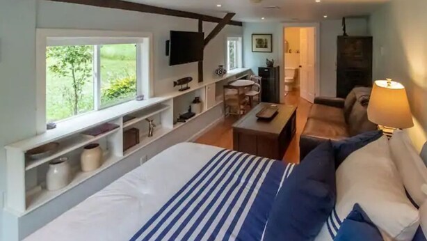 A bedroom with a bed, a TV mounted on the wall, a window with a view of greenery, and a small seating area with a table and chairs.