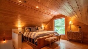 A cozy attic bedroom with a wooden ceiling, a large window, a bed with a striped blanket, a bedside table with a lamp, and a cabinet. - The Loft At The Inn At High Falls (High Falls)