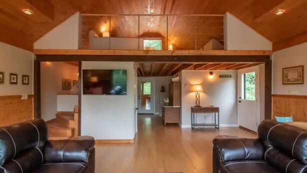 A spacious living area with a high ceiling, wooden beams, two leather sofas, a central rug, and a staircase leading to an upper level. - The Loft At The Inn At High Falls (High Falls)