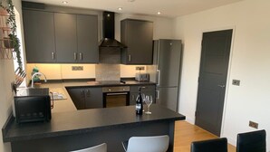 A modern kitchen with dark cabinetry, a central island with stools, a dining table with chairs, and a refrigerator. - The Mariners' Beacon Torquay (Torquay)