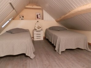 Une petite chambre mansardée triangulaire, avec deux lits simples, une petite commode et un vase décoratif.