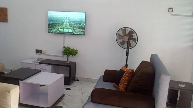 A modern living room with a sofa, a coffee table, a TV mounted on the wall, and a standing fan. - Spacious Serviced Apartments in Owerri,Accommodation Corporate and Private Stays (Owerri)