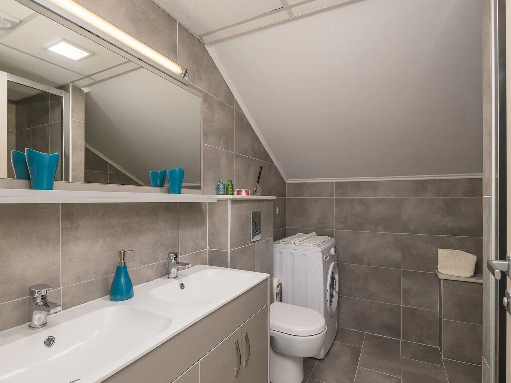 A modern bathroom with a white sink, a toilet, and a washing machine. There are shelves with toiletries and a mirror above the sink.