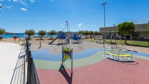 Un parque infantil con toboganes y estructuras de escalada, un área vallada y un edificio con la inscripción 'ROOFTOP BAR' al fondo.