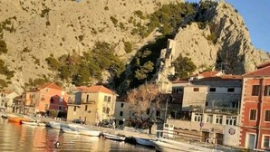 Un muelle con barcos amarrados, edificios con techos rojos y una cadena montañosa al fondo.