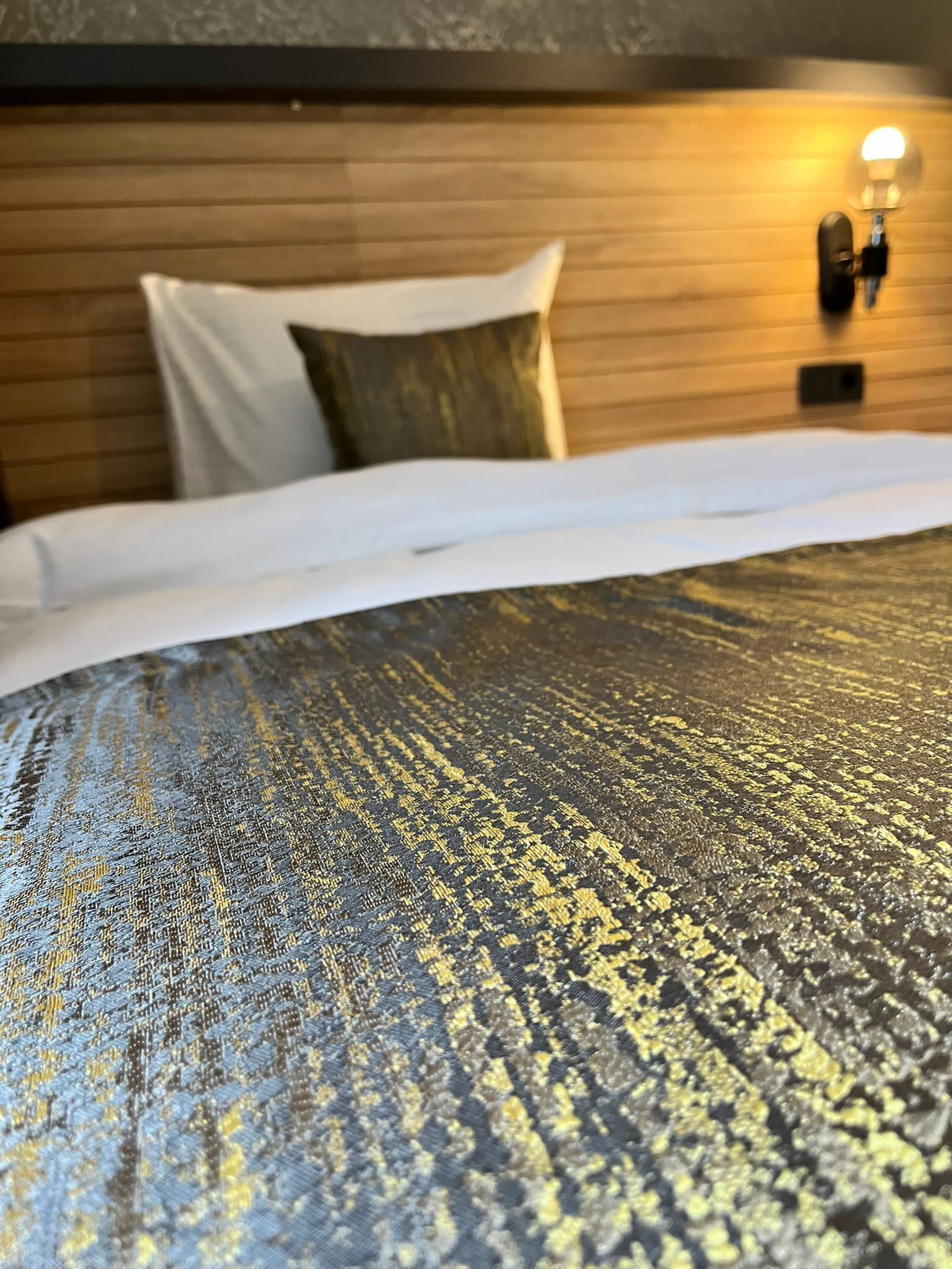 Una cama con una colcha dorada y negra de textura, un cabecero de madera y una lámpara fijada a la pared.