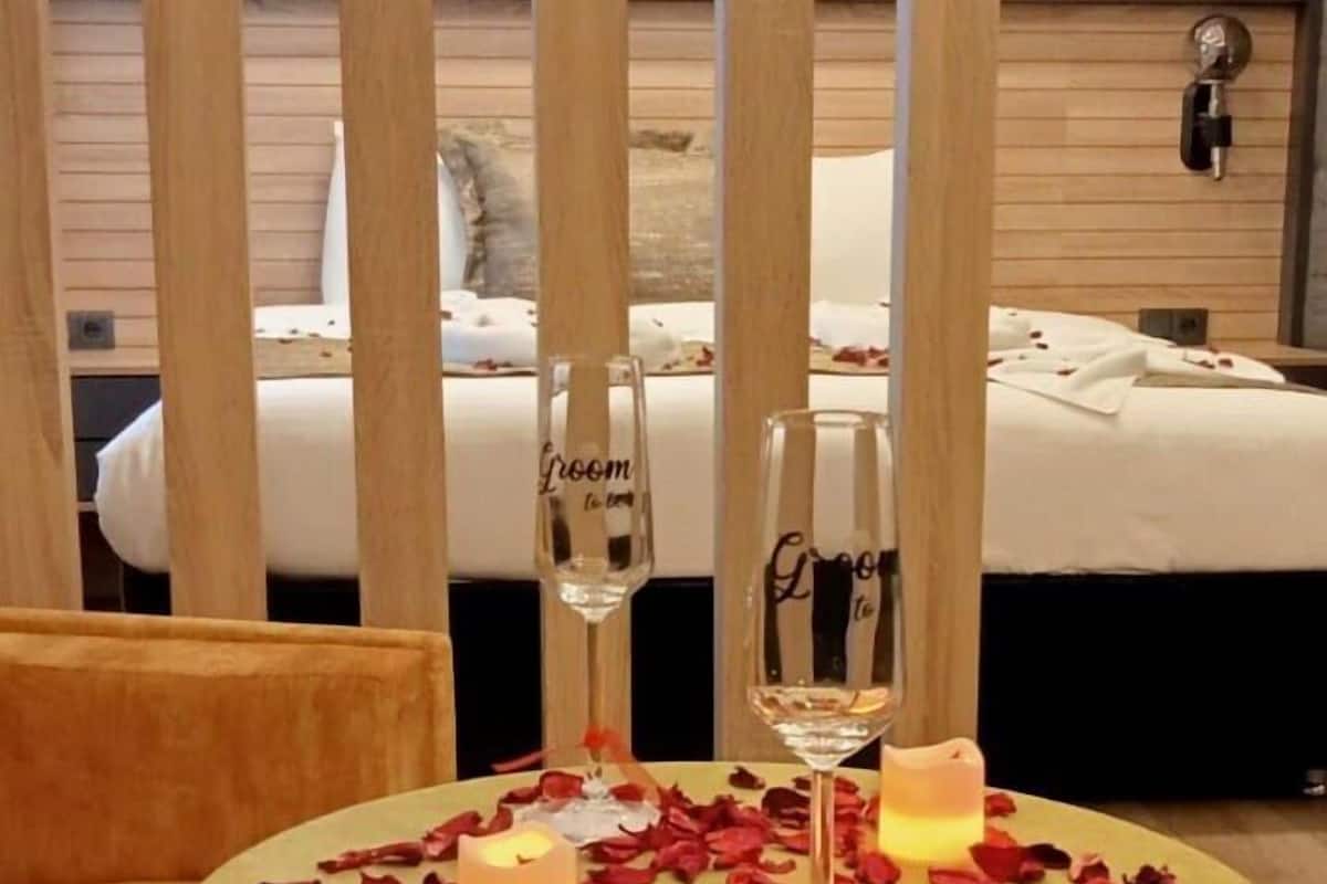 A hotel room with a bed, a table with rose petals, candles, and two glasses labeled "Groom" and "Bride".